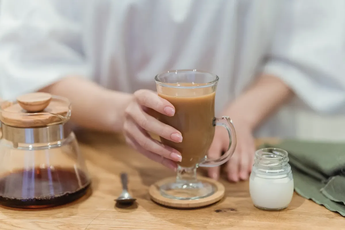 A close-up of a hand holding a coffee mug on a wooden table, with a pot and milk jar.
